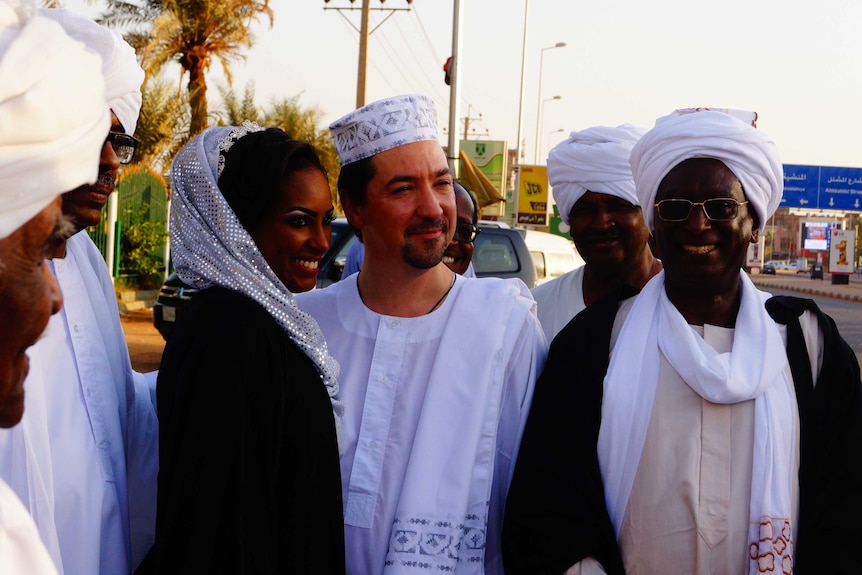 A group of people in white traditional Sudanese dress standing by the roadside and smiling.