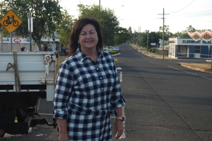 A woman with long brown hair, wearing a checked black and white dress, stands smiling next to a country road.