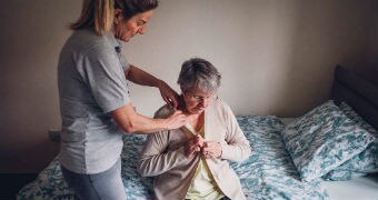 An aged care working standing up helps dress a senior woman sitting on her bed.