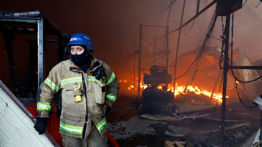 A rescue worker wearing a helmet standing and looking out from a damaged warehouse in which there is fire and twisted metal.