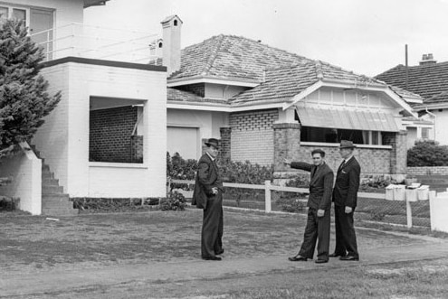 Black and white pic of serial killer Eric Edgar Cooke pointing with detectives outside a house.