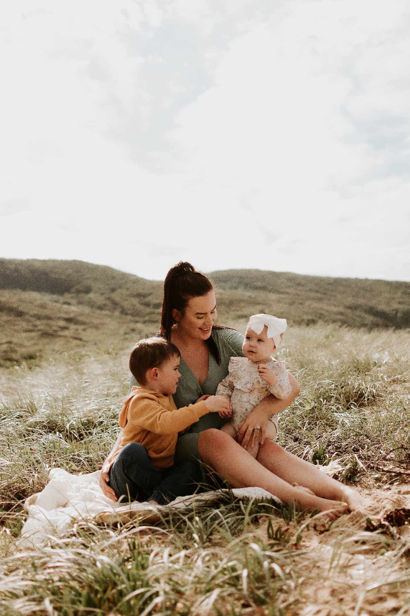 A woman sitting on the grass, cradling her two children, a toddler boy and a baby girl