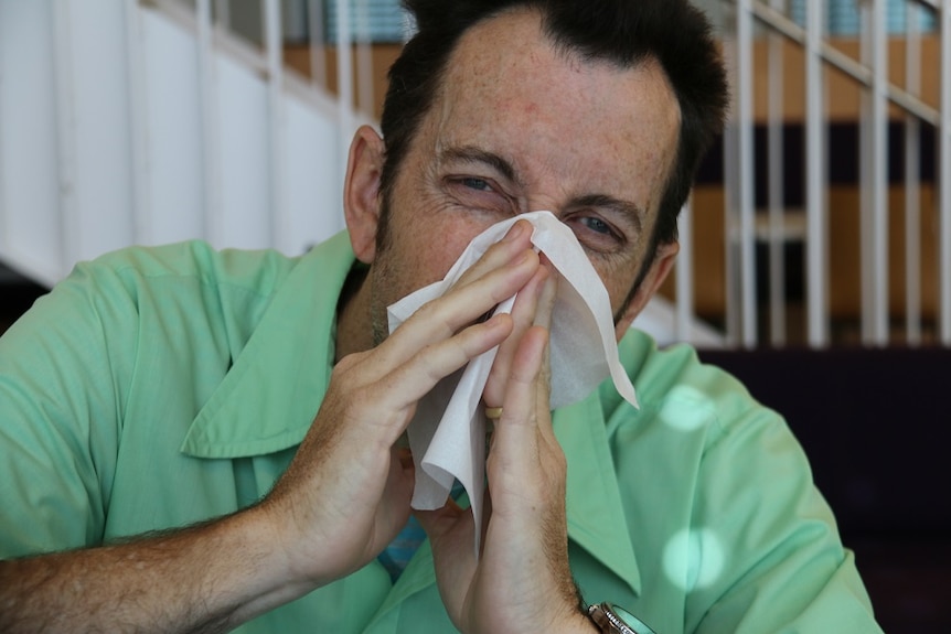 A man in a green shirt blows his nose with a tissue.