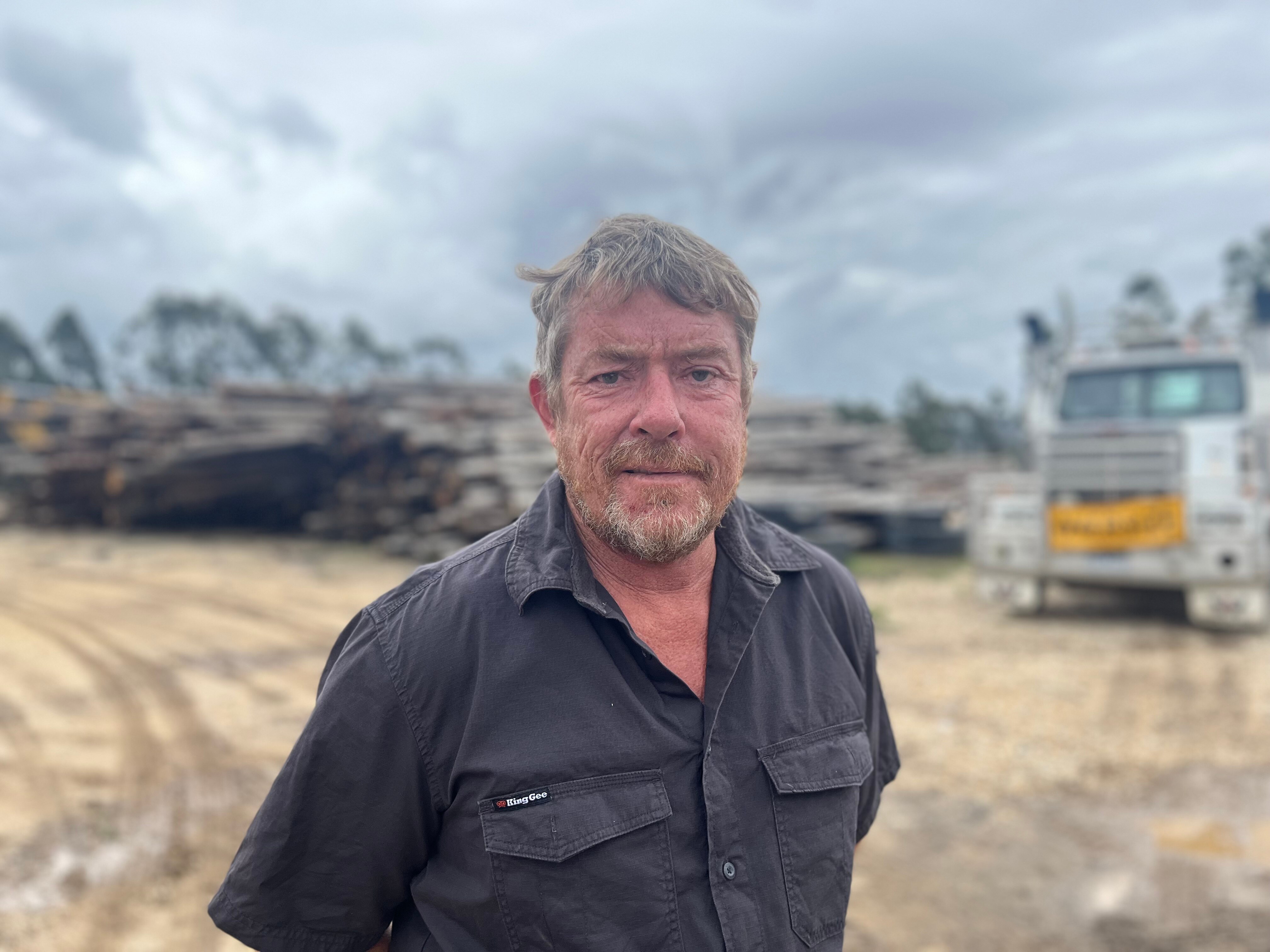 Man standing at sawmill site with logs and truck in background