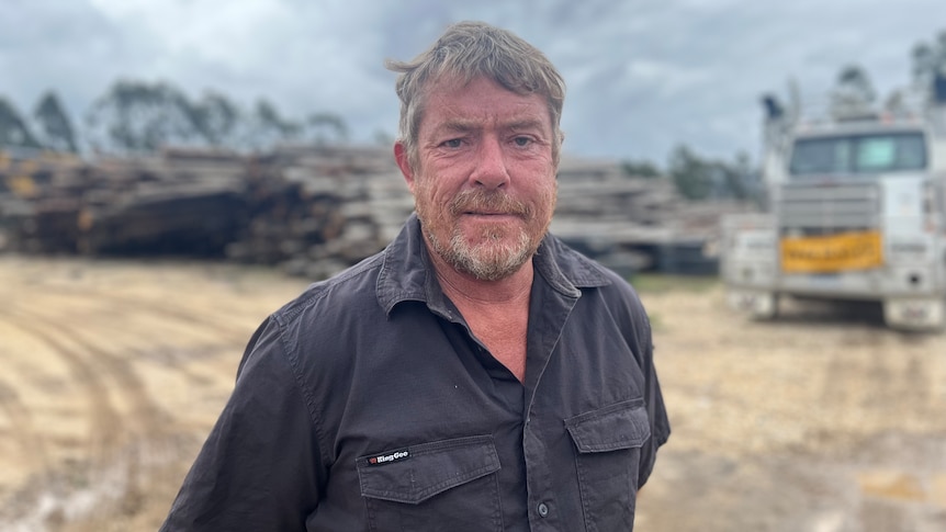 Man standing at sawmill site with logs and truck in background