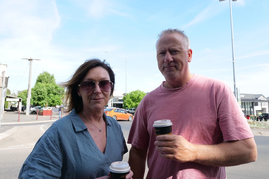 A man and a woman, both middle-aged, look solemn as they stand holding takeway coffees on a road in a country town.