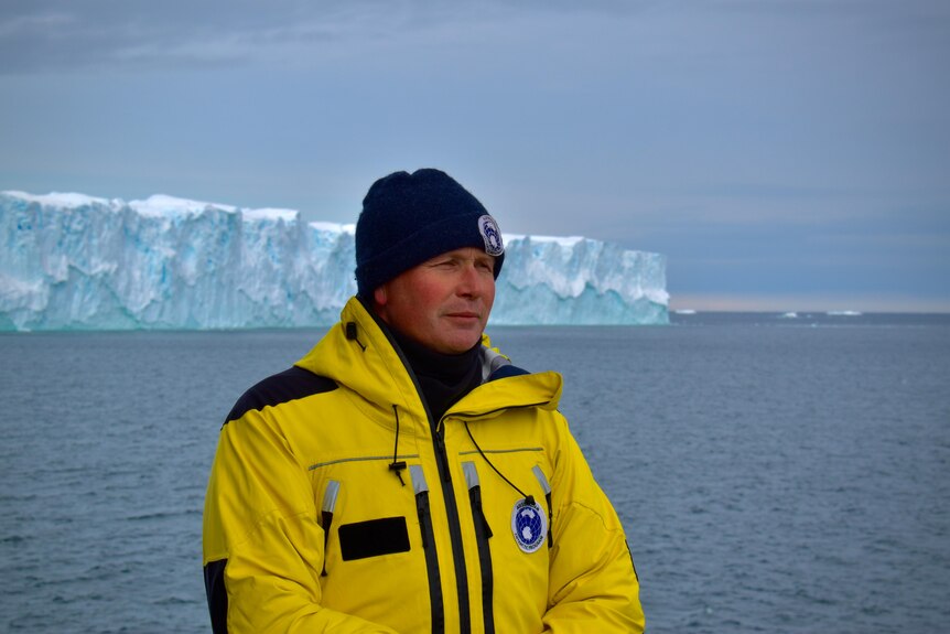 A man in a heavy weatherproof jacket stands on the deck of a ship, a glacier visible behind him.