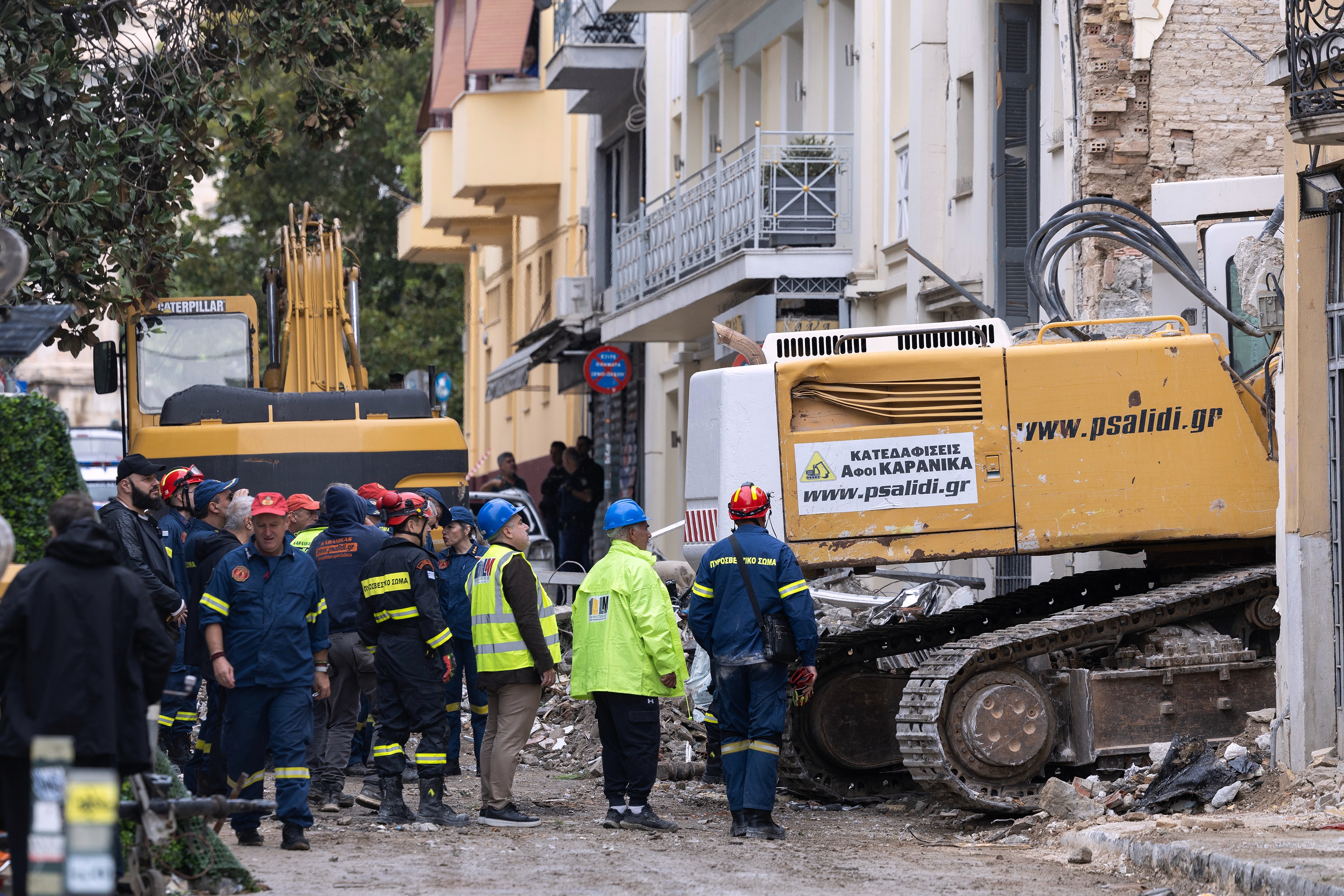 Rescue workers in blue, black and bright yellow overalls and clothing, wearing hard hats in a street next to a yellow digger.