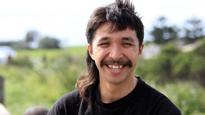 A youngish man with a black moustache smiles while wearing a black t-shirt near the beach.