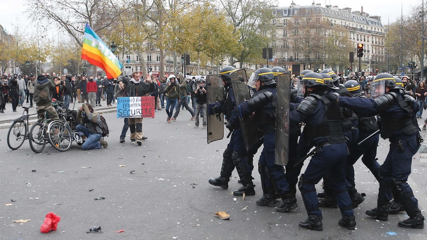 Paris protesters take photos and hide from police marching forward with riot gear on a dirty street.