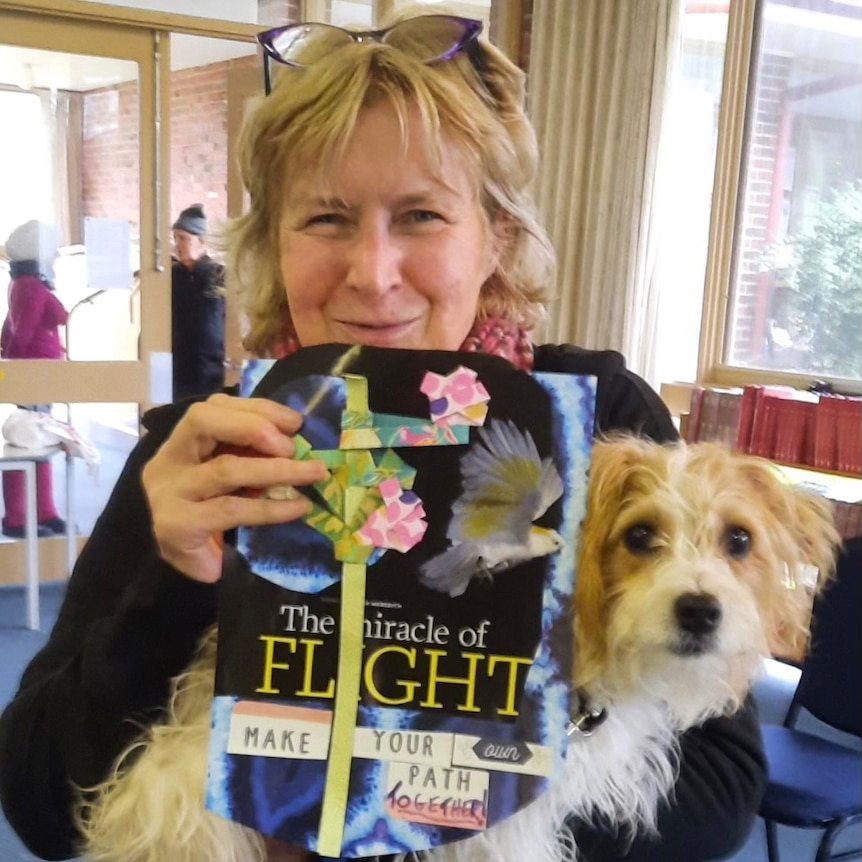 A woman holds a small dog and a book, titled 'The Miracle of Flight', while smiling.