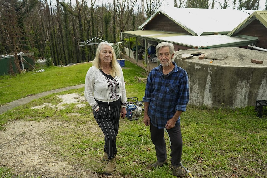 A couple stand in front of their house and regrowing burned trees.