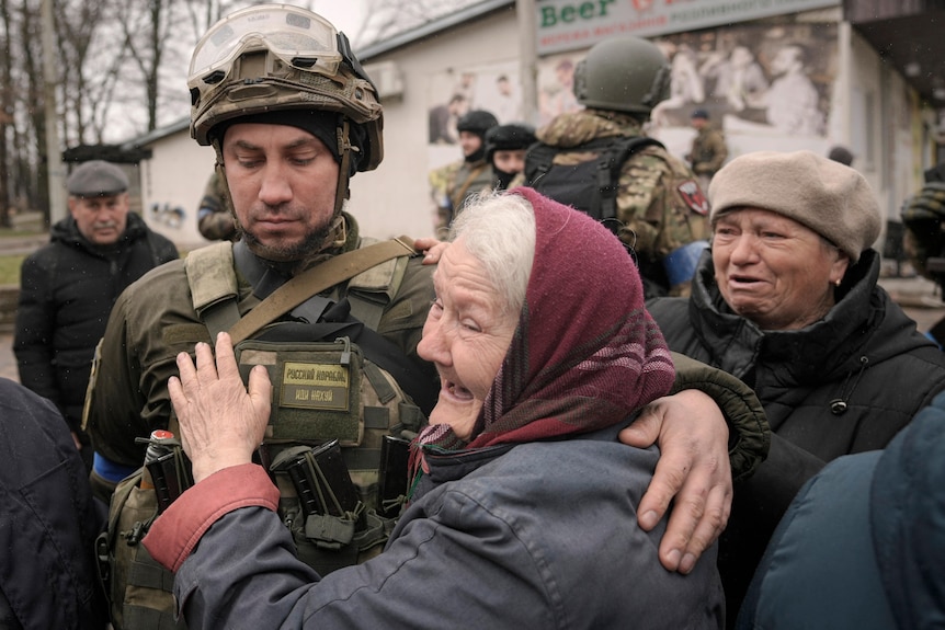 An elderly woman in a headscarf smiles as she hugs a soldier with a downcast look on his face.