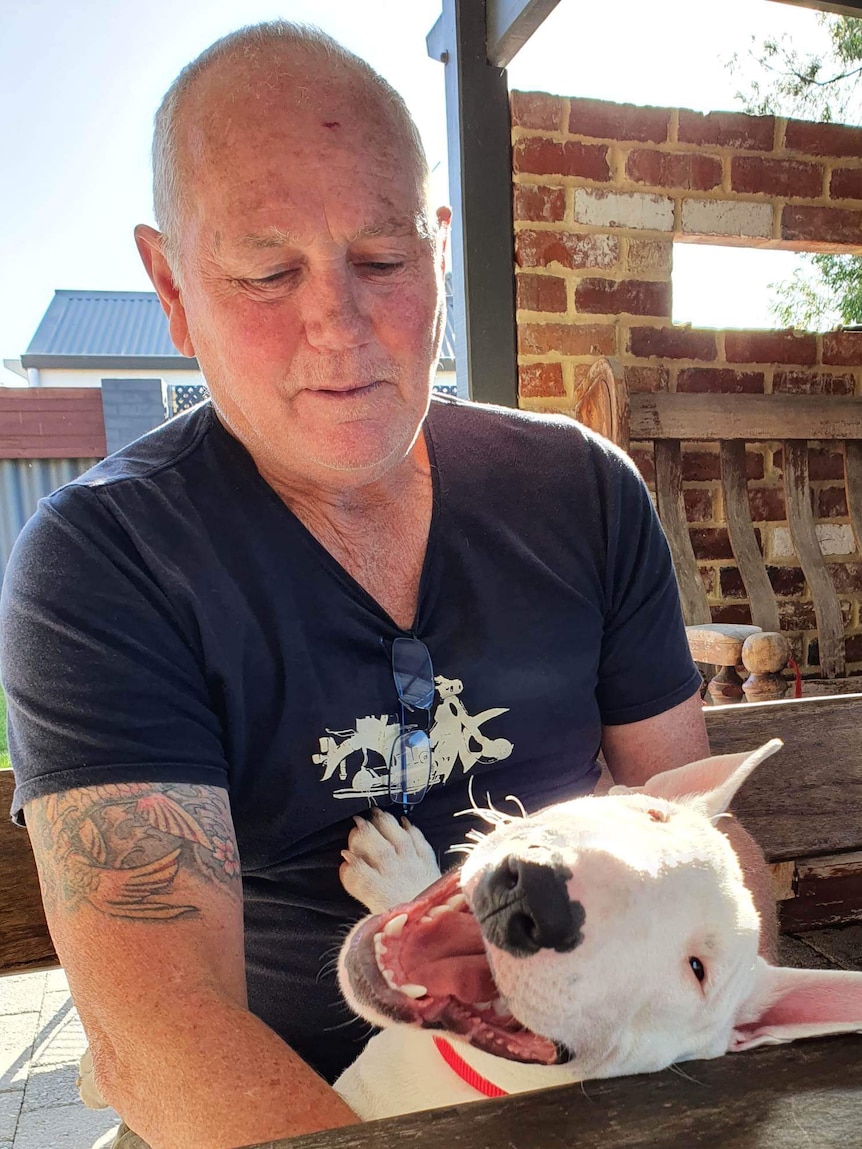 A man with a bald head and tattoos sits outside, he is cradling a dog.