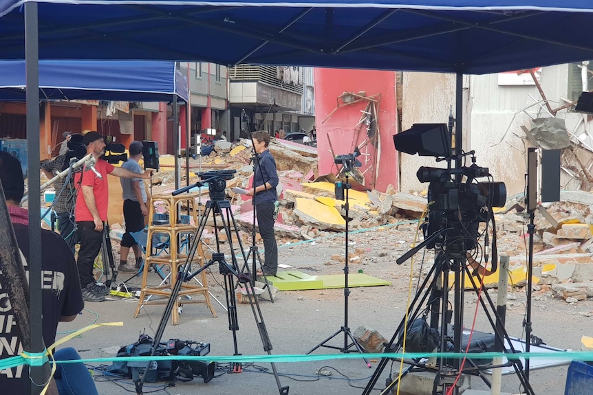 Journalist and cameraman standing with equipment in front of damaged buildings.