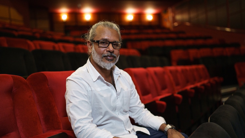 47-year-old Sri Lankan man with grey beard, moustache and long hair pulled back, wearing a white shirt, sitting in theatre.