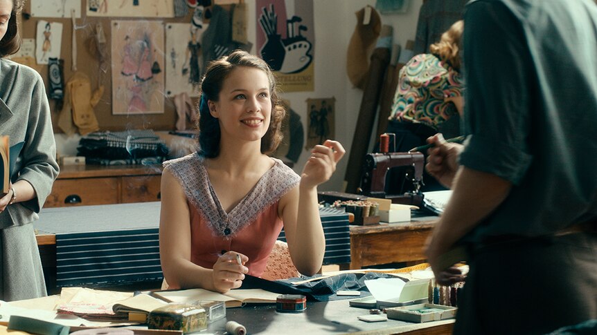 Colour still of Paula Beer smoking, seated at desk and smiling at man in 2018 film Never Look Away.