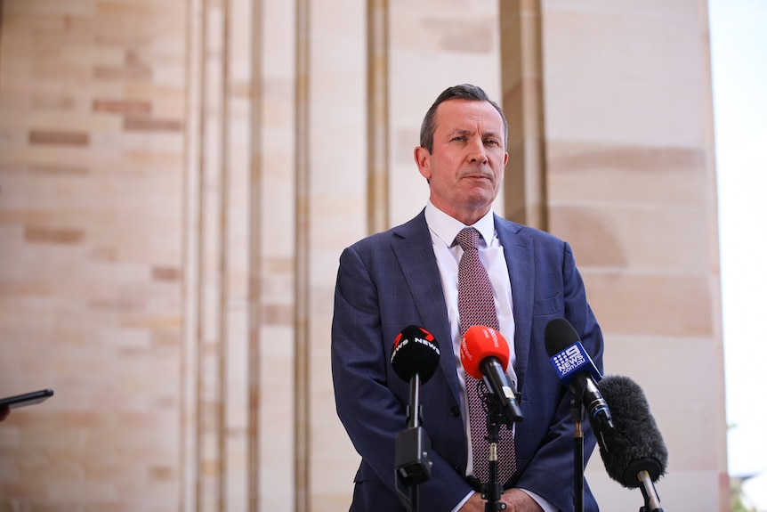 A man wearing a suit stands in front of microphones looking serious.