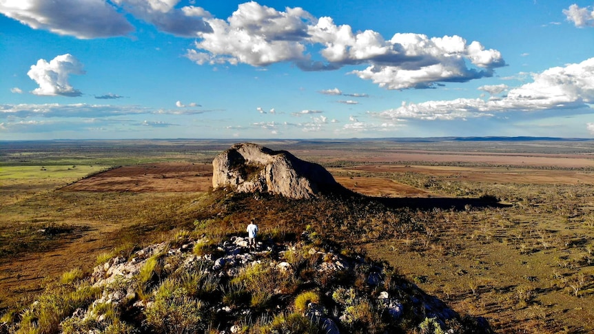 Mountain range jutting out of the ground, a man stands atop in the foreground, blue sky behind.