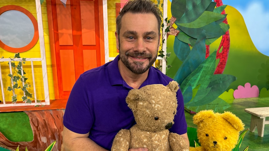 A man holds a teddy bear in front of a colourful house on a television set. 