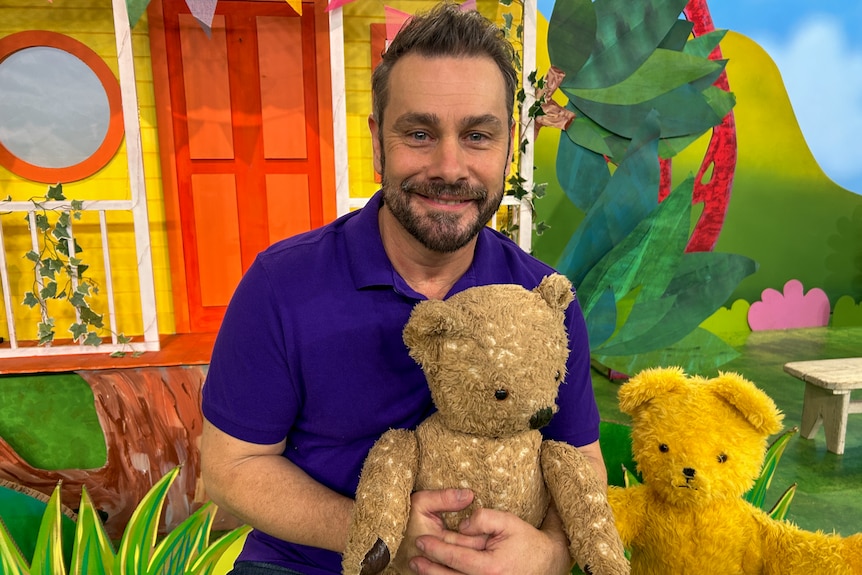 A man holds a teddy bear in front of a colourful house on a television set. 