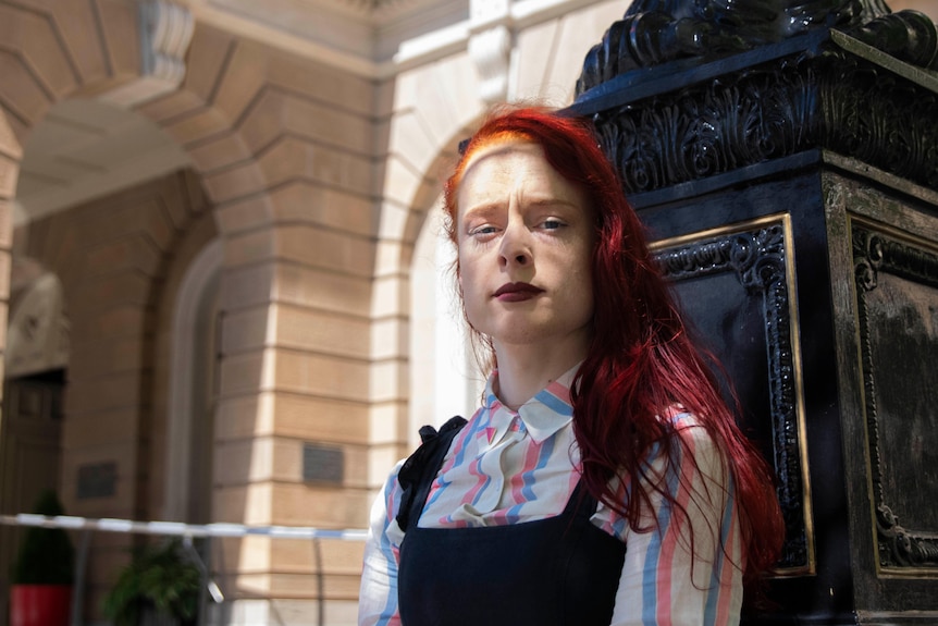 woman with orange and red hair and a brightly coloured shirt stands outside a sandstone building 