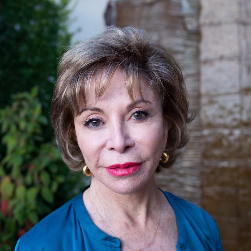 Isabel Allende from shoulders up. She's standing in front of a plant covered wall.