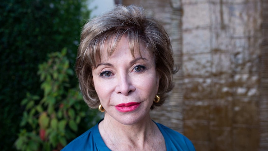 Isabel Allende from shoulders up. She's standing in front of a plant covered wall.