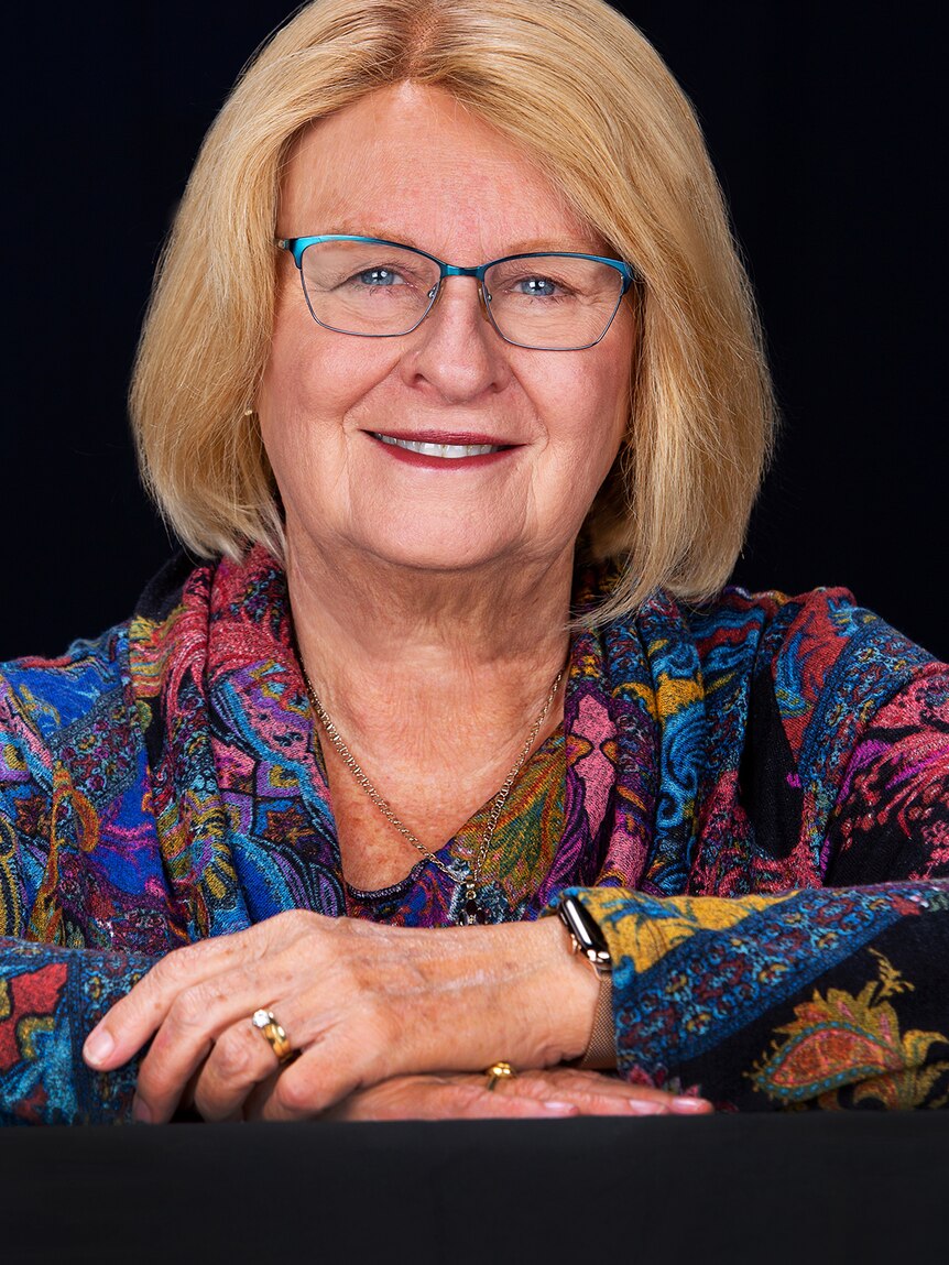 A profile shot of an older, bespectacled woman with blonde hair, smiling.