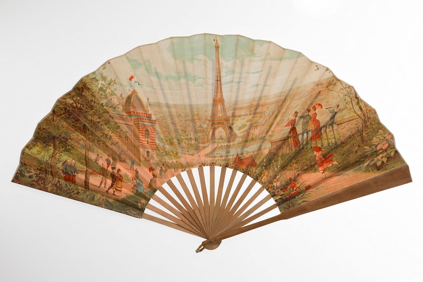 A hand-held fan spread open to reveal a panoramic image of Paris, the Eiffel Tower and people wearing attire from the 1880s.