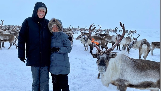 A couple wearing snow jackets with hoods stands on the snow with a reindeer herd behind them.