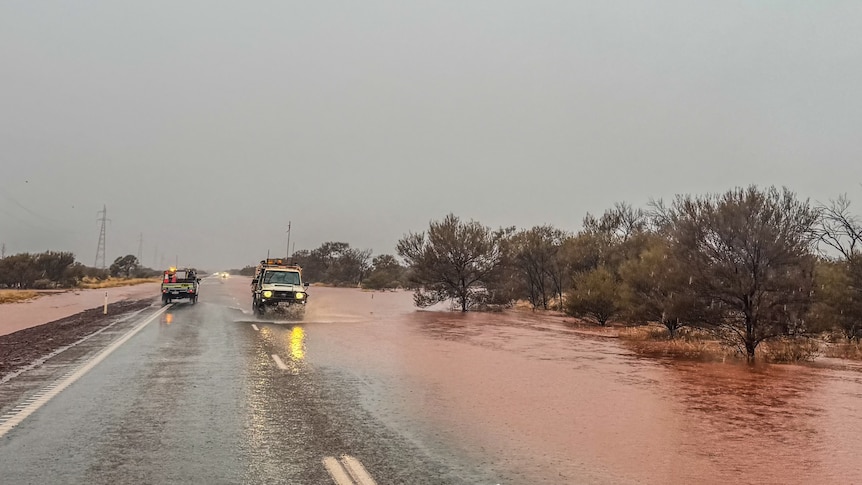 Cars travel along a flooded road with grey skies, and flooded bushland on the right.