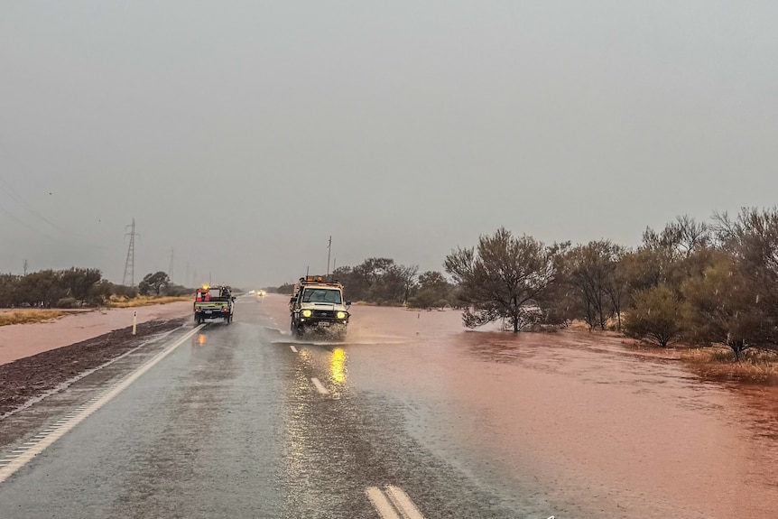 Cars travel along a flooded road with grey skies, and flooded bushland on the right.