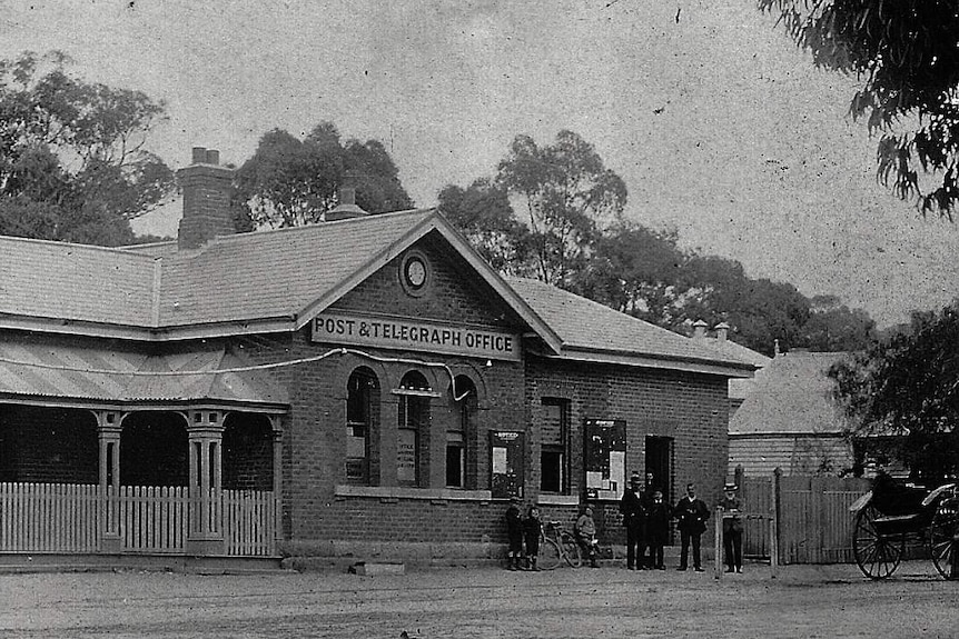 Black and white photo of single storey brick post office. A few people gathered out the front next to a horse and cart.