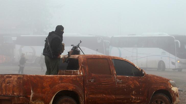 A rebel fighter stands with a gun on a vehicle near buses carrying people.