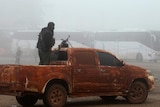 A rebel fighter stands with a gun on a vehicle near buses carrying people.
