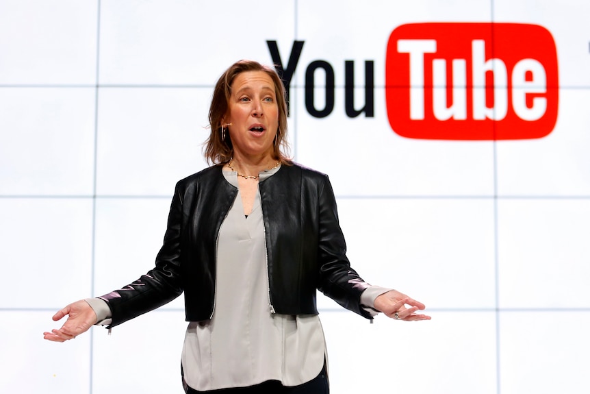 A woman stands speaking in front of a large YouTube logo illuminated behind her. 