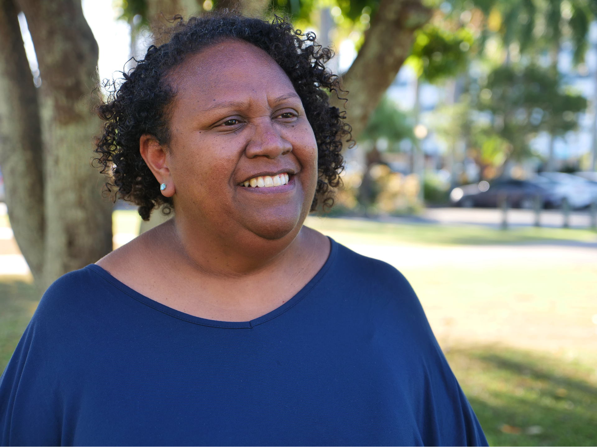 a woman with a blue top on stands in a park, smiling off into the distance