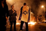 An Israeli protester draped in an Israeli flag and others gather on a street around burning objects.