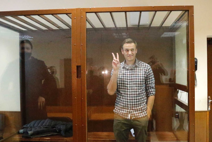 A man gives a peace sign with his right hand as he stands in a glass box dock inside a court with two guards nearby