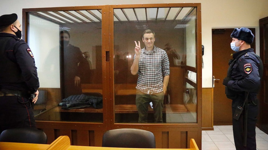 A man gives a peace sign with his right hand as he stands in a glass box dock inside a court with two guards nearby