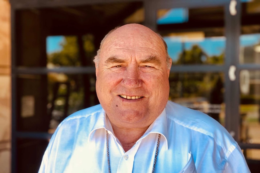 A close up shot of large balding man wearing a blue shirt and chain around his neck.