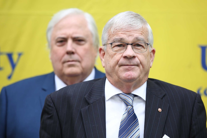 Brian Burston stares tight-lipped into the camera while Clive Palmer stands behind with a stern expression on his face.