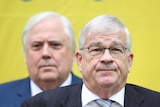 Brian Burston stares tight-lipped into the camera while Clive Palmer stands behind with a stern expression on his face.