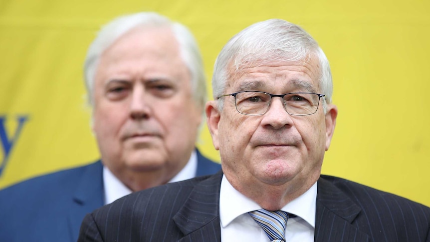 Brian Burston stares tight-lipped into the camera while Clive Palmer stands behind with a stern expression on his face.