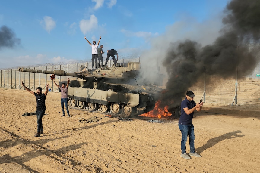 Palestinians celebrate on top of and nearby a Israeli tank which has been set on fire near a fence