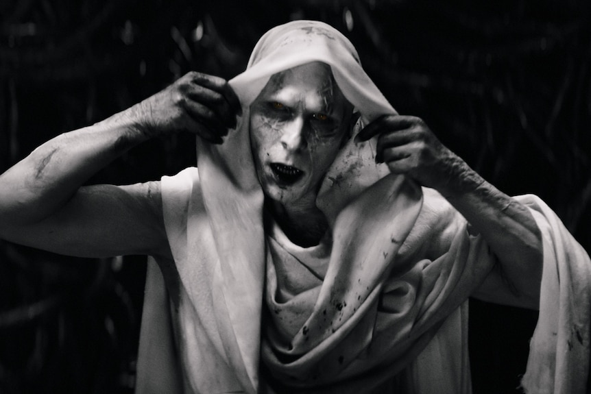 Actor in white make-up with darkened eyes, drawing up a white hood over his head.