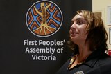 A woman standing in front of a sign that reads First Peoples' Assembly of Victoria