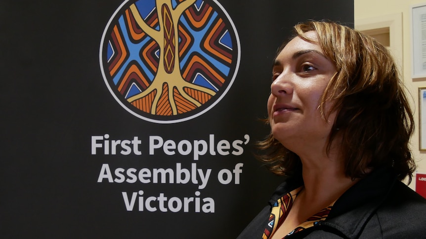 A woman standing in front of a sign that reads First Peoples' Assembly of Victoria