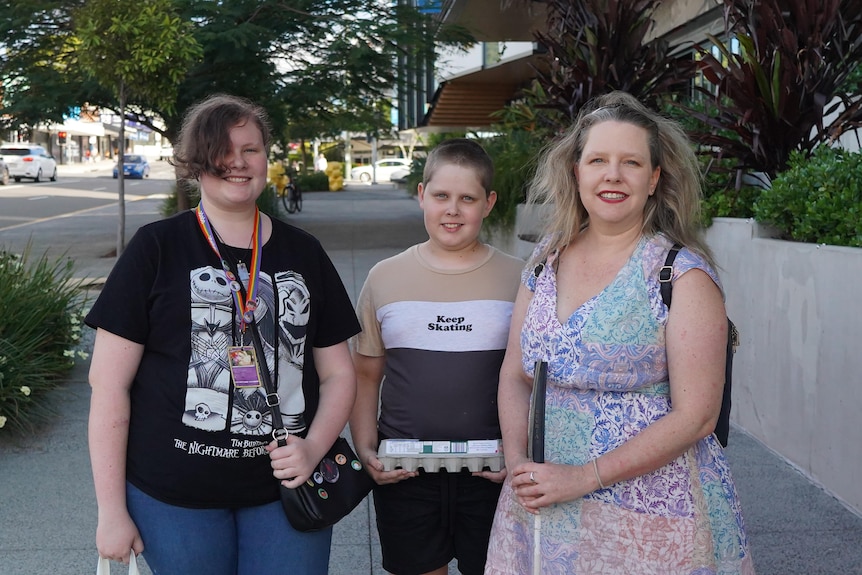 Sarah Evans is holding a cane, smiling and standing next to her two children outdoors.